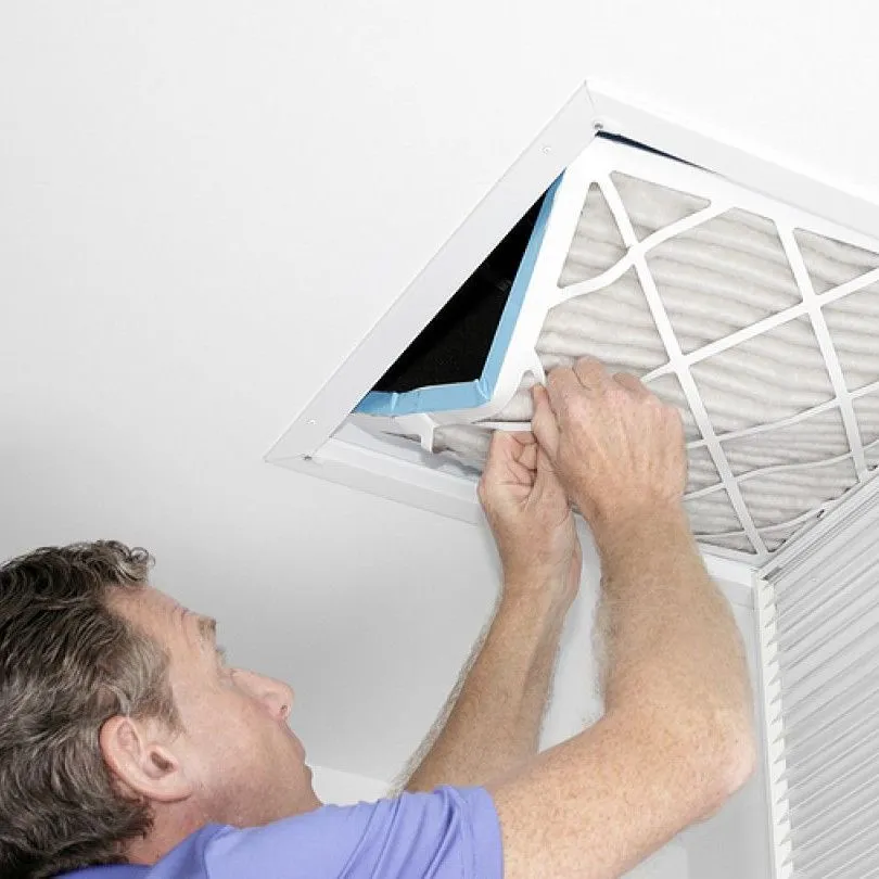 A person in a purple shirt replaces a pleated air filter inside a ceiling-mounted HVAC return vent.